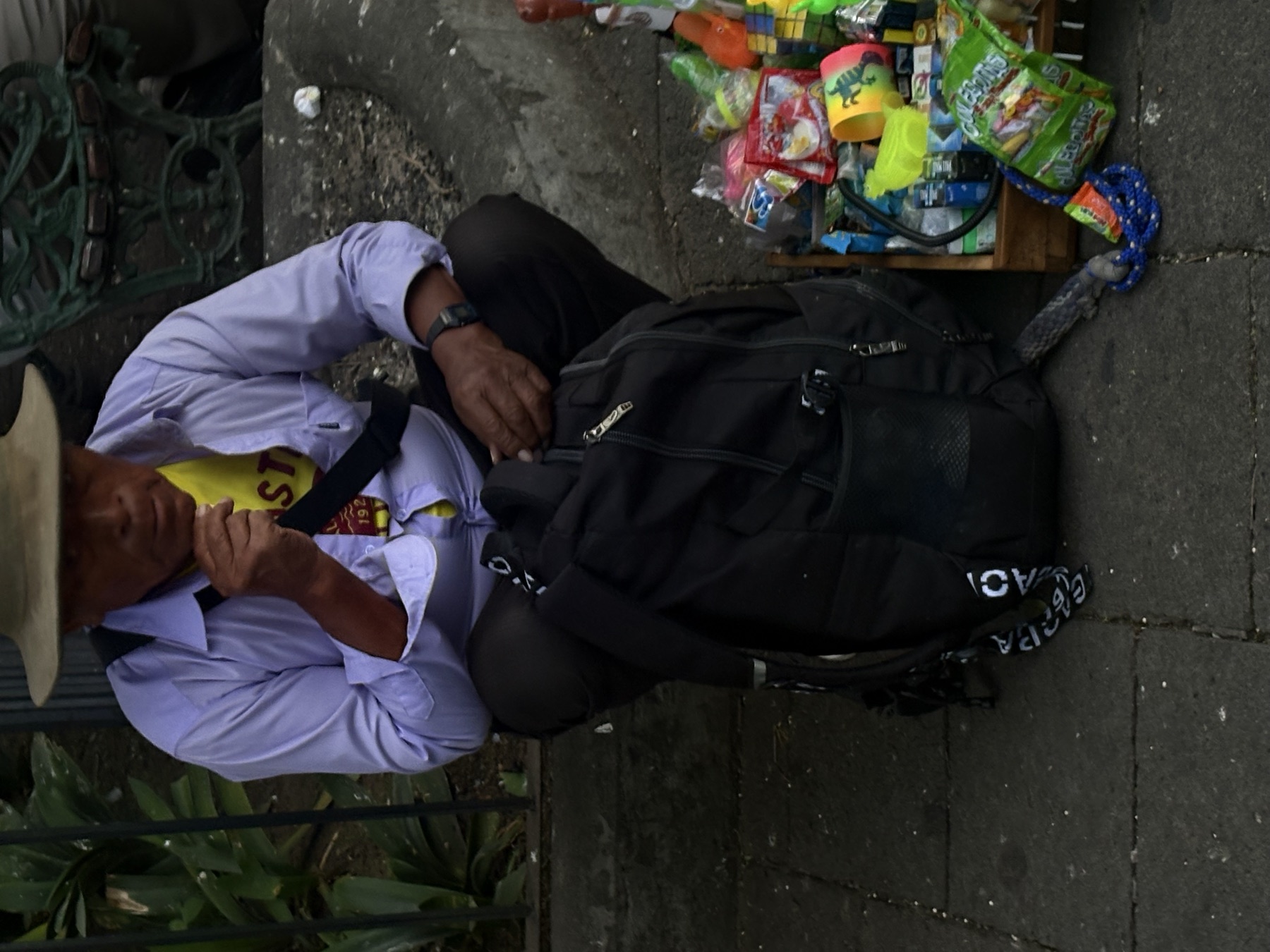 Man in a straw hat eating on a kerb next to a backpack